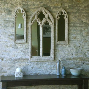 May include: Three arched mirrors with ornate frames are mounted on a stone wall. The mirrors are different sizes and are arranged in a row. A dark wooden table with a candle and two bottles sits in front of the mirrors.