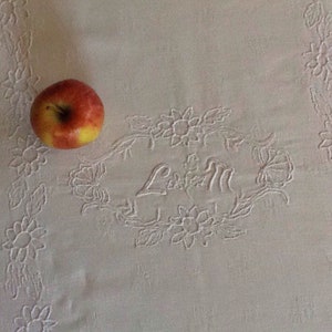 Op de afbeelding: Een wit tafelkleed met geborduurde bloemenmotieven en de naam "L.E.M." in cursief schrift. Een rode en gele appel ligt op het tafelkleed.