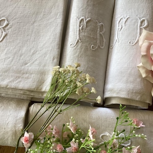 May include: A set of off-white linen napkins featuring white embroidered monograms with the letters FB. The napkins are folded and stacked, accompanied by a sprig of small white flowers and a large, pale pink rose blossom on the right side.
