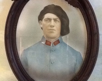 Pode incluir: Uma moldura de retrato oval vintage com acabamento em madeira escura. A moldura apresenta talhas ornamentadas e um inserto oval de vidro. O retrato no interior mostra um homem vestindo um uniforme azul e um boné escuro.