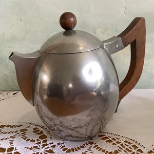 May include: A silver teapot with a wooden handle and a wooden knob on the lid. The teapot is sitting on a white lace tablecloth.