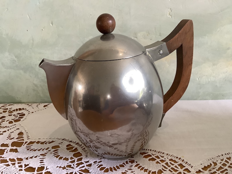 May include: A silver teapot with a wooden handle and a wooden knob on the lid. The teapot is sitting on a white lace tablecloth.