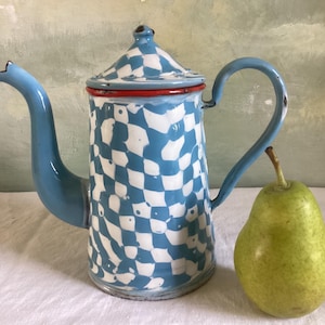 Puede incluir: Una cafetera vintage de esmalte con un patrón de cuadros azules y blancos. La cafetera tiene un asa y un pico de color azul claro, con un borde rojo. Una pera verde está al lado de la cafetera.