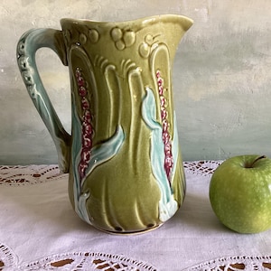 May include: A green ceramic pitcher with a floral design. The pitcher has a handle and a spout. The floral design is in shades of green, blue, and pink.