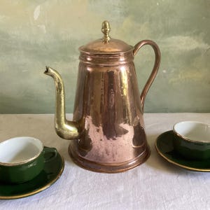 May include: A polished copper coffee pot with a brass spout, handle, and lid finial, accompanied by two dark green ceramic teacups with white interiors and gold-toned rims, each resting on a matching dark green saucer with a gold-toned edge.