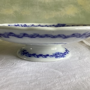 Può includere: Una ciotola bianca con piedistallo con un motivo floreale blu e bianco. La ciotola è decorata con un bordo blu e bianco.