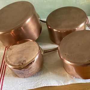 May include: Four copper saucepans with rounded sides and long handles are arranged on a white and red striped cloth. The pans have a shiny, reflective surface, and are of varying sizes, suggesting they are a set for cooking.