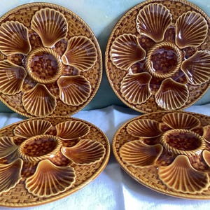 May include: Four round ceramic oyster plates in a glossy brown glaze. Each plate features six shell-shaped indentations arranged around a central circular well, with a textured basket-weave pattern covering the remaining surface area.