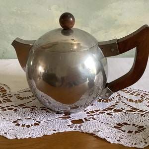 May include: A silver teapot with a wooden handle and a wooden knob on the lid. The teapot is sitting on a white lace tablecloth.