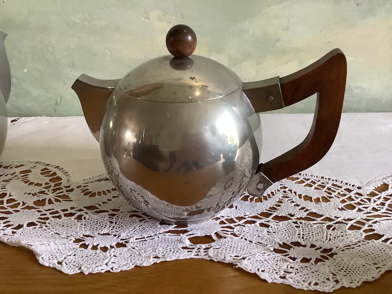 May include: A silver teapot with a wooden handle and a wooden knob on the lid. The teapot is sitting on a white lace tablecloth.