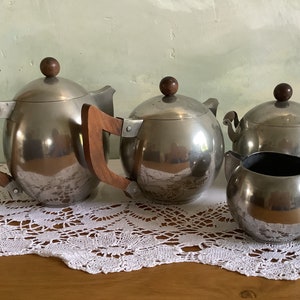 May include: A silver tea set with wooden handles. The set includes a teapot, creamer, and sugar bowl. The tea set is on a white lace tablecloth.