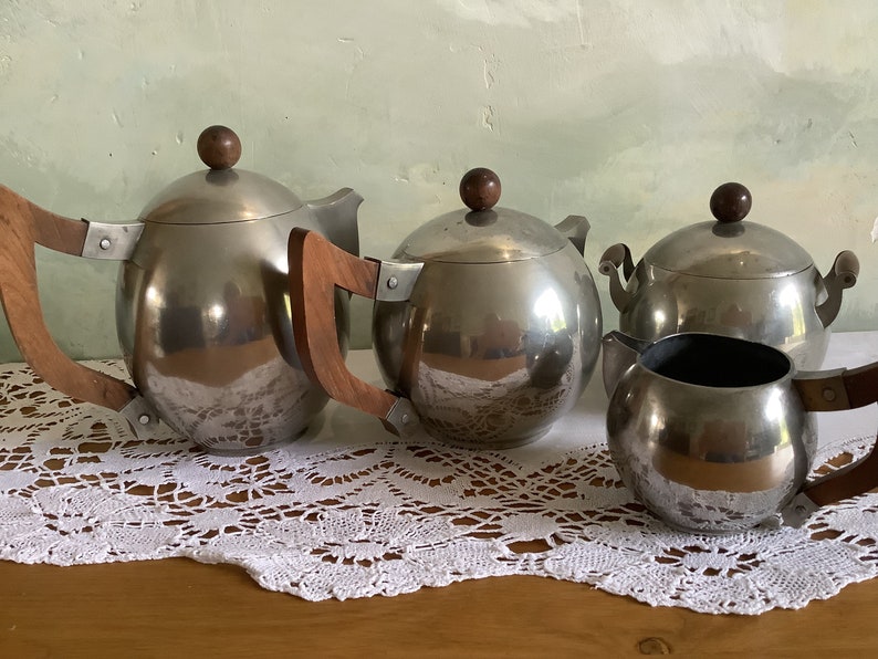 May include: A silver tea set with wooden handles. The set includes a teapot, creamer, and sugar bowl. The tea set is on a white lace tablecloth.