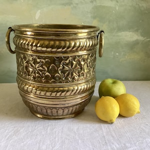 May include: A polished brass bucket featuring two side handles and a decorative embossed pattern of stars and geometric shapes. The bucket has a wide rim and a rounded base, accompanied by one green apple and two yellow lemons placed nearby.