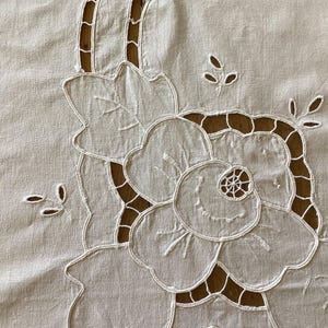 以下が含まれることがあります： 明るい色の生地に施された、白い刺繍の花柄のクローズアップ。 詳細な花びらと切り抜かれた中心部を持つ大きな花が特徴で、葉や小さな花のアクセントで囲まれています。 背景は対照的な茶色です。