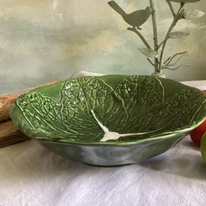 Peut inclure: Un saladier en céramique verte en forme de feuille de chou, avec des nervures détaillées et une finition brillante. Un poivron rouge, une tomate et une pomme verte sont à côté du saladier. Une baguette est sur une planche de bois.