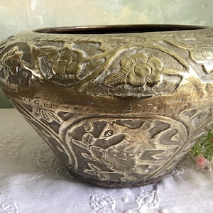 Op de afbeelding: Een grote, antieke messing kom met ingewikkelde bloemen- en vogelreliëfsnijwerken. De kom heeft een brede, afgeronde vorm en een donker interieur. De buitenkant is een mix van goud- en zilverkleuren, met gedetailleerde patronen. Een decoratief stuk.