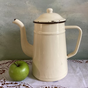 May include: Cream-colored vintage enamelware coffee pot with a long spout and handle. The pot has a dark brown trim and a lid with a small knob. A green apple sits next to the pot on a white lace tablecloth.