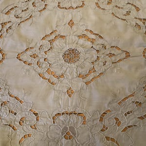 Pode incluir: Toalha de mesa bordada creme com um design floral. A toalha de mesa apresenta grandes padrões florais vazados nas bordas e um medalhão floral central. O fundo é de cor castanho claro, criando um contraste com o bordado branco.