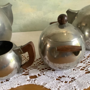 May include: A silver tea set with a teapot, creamer, and sugar bowl. The teapot and sugar bowl have wooden handles. The set is on a white lace tablecloth.