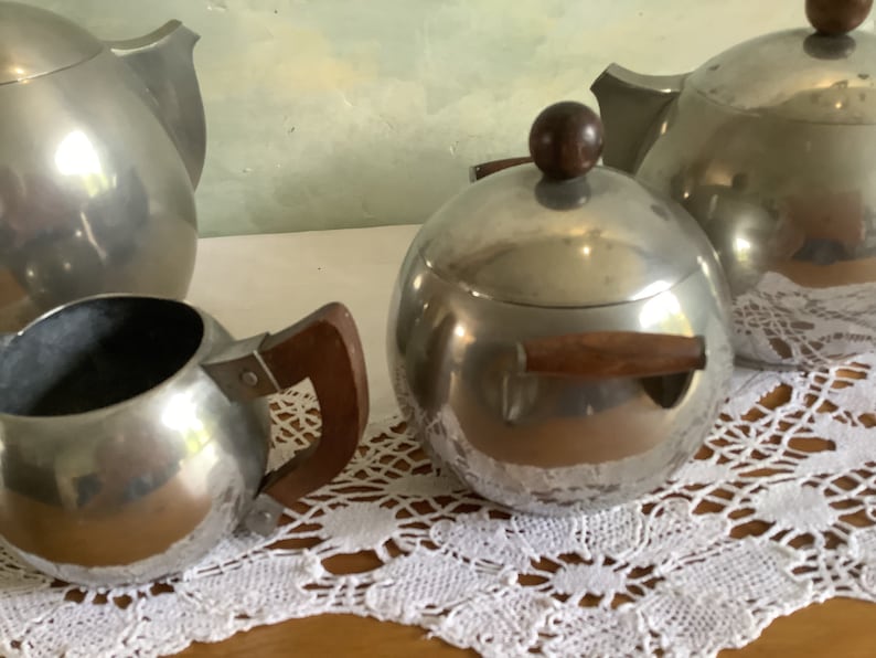 May include: A silver tea set with a teapot, creamer, and sugar bowl. The teapot and sugar bowl have wooden handles. The set is on a white lace tablecloth.