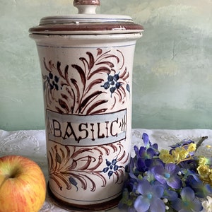 Puede incluir: Tarro de cerámica blanca con tapa marrón y diseño floral azul y marrón. El tarro está etiquetado con "BASILIC" en letras azules.