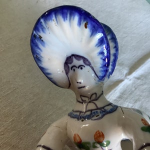 Può includere: Una statuetta in ceramica di una donna con un cappello blu e bianco. La donna ha un viso dipinto con capelli castani e un vestito bianco con dettagli floreali.