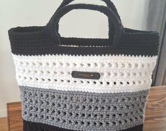 Puede incluir: Un bolso tote de ganchillo con rayas horizontales en negro, blanco y gris. El bolso tiene dos asas negras y una pequeña etiqueta rectangular de color marrón oscuro centrada en la sección blanca con el texto Hand Made with love en dorado.