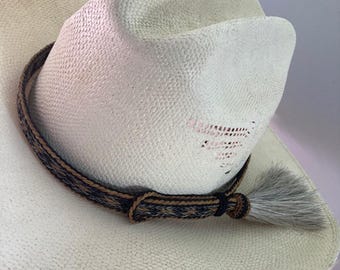 Op de afbeelding: Een crèmekleurige cowboyhoed met een decoratieve band met een geweven patroon in bruin- en marineblauw. Een grijze kwast hangt aan de zijkant. De hoed is gemaakt van geweven materiaal.