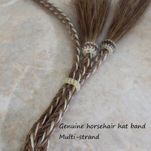 May include: A brown and white braided horsehair hat band with double-sided tassels. The hat band is made of multiple strands of horsehair and is tied with a knot at the end. The text "Genuine horsehair hat band Multi-strand Double side tassels from Knot-a-tail.com" is visible in the image.