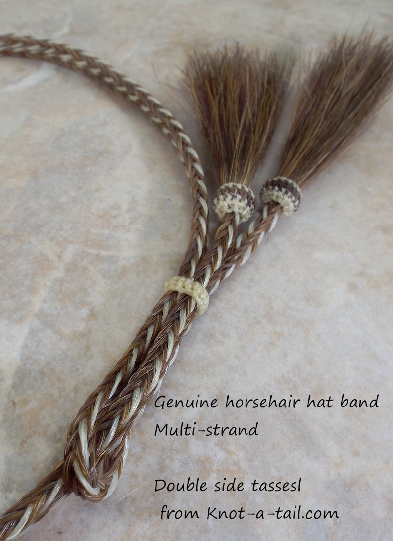 May include: A brown and white braided horsehair hat band with double-sided tassels. The hat band is made of multiple strands of horsehair and is tied with a knot at the end. The text "Genuine horsehair hat band Multi-strand Double side tassels from Knot-a-tail.com" is visible in the image.