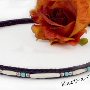 May include: A black braided leather cord with turquoise and white beads, tied into a loop with a black tassel. The cord is decorated with silver beads. The text "Knot-a-Tail.com" is visible in the bottom right corner of the image.