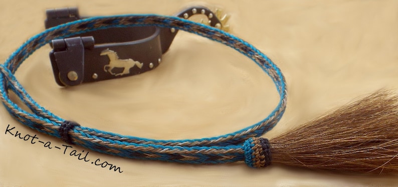 May include: A turquoise, brown, and beige braided leather horse halter with a brown horsehair tassel. The halter has a silver buckle with a horse silhouette.