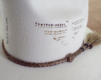 May include: A white straw hat with a decorative woven band in brown and black. The hat features a decorative cut-out design and a tassel. The text "knot-a-tail.com" is visible on the side of the hat.