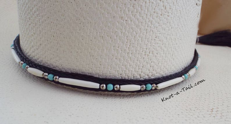 May include: A white cowboy hat with a black braided hat band accented with white and turquoise beads. The hat band has the text "Knot-a-Tail.com" on it.
