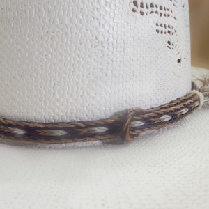 Puede incluir: Primer plano de un sombrero de vaquero blanco con una banda trenzada decorativa. La banda presenta un patr&oacute;n de hebras marrones, negras y blancas. Una peque&ntilde;a borla cuelga del lateral del sombrero.