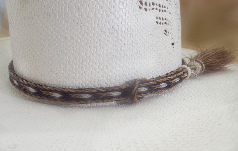 Puede incluir: Primer plano de un sombrero de vaquero blanco con una banda trenzada decorativa. La banda presenta un patr&oacute;n de hebras marrones, negras y blancas. Una peque&ntilde;a borla cuelga del lateral del sombrero.