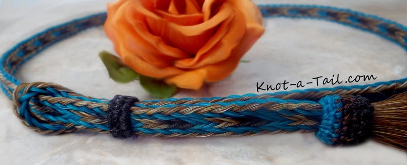 May include: A turquoise, brown, and black braided horsehair rope halter with a brown horsehair tassel. The halter is braided with a knot pattern and has a decorative knot at the end of the tassel. Knot-a-Tail.com is printed on the halter.