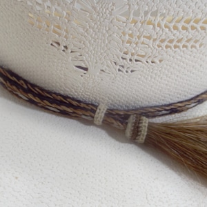Puede incluir: Primer plano de un sombrero de vaquero color crema con una banda trenzada decorativa en marr&oacute;n y beige. Una borla a juego cuelga del lateral, a&ntilde;adiendo un toque r&uacute;stico al dise&ntilde;o del sombrero.
