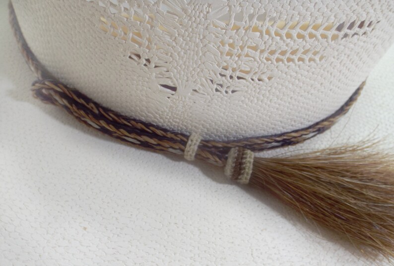Puede incluir: Primer plano de un sombrero de vaquero color crema con una banda trenzada decorativa en marr&oacute;n y beige. Una borla a juego cuelga del lateral, a&ntilde;adiendo un toque r&uacute;stico al dise&ntilde;o del sombrero.