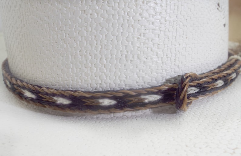 Puede incluir: Primer plano de un sombrero de vaquero blanco con una cinta tejida. La cinta es una mezcla de fibras marrones, negras y blancas, creando un dise&ntilde;o estampado. El sombrero tiene una superficie texturizada.
