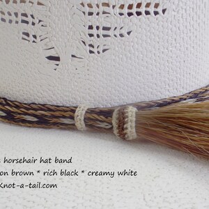 Puede incluir: Primer plano de un sombrero de vaquero blanco con una banda decorativa de crin de caballo trenzada. La banda presenta colores marr&oacute;n canela, negro intenso y blanco crema. Una borla de crin de caballo cuelga de la banda.