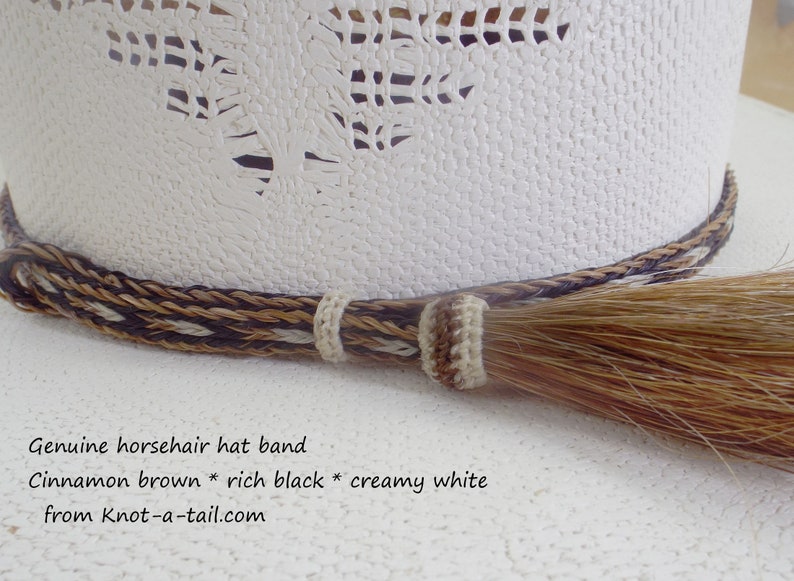 Puede incluir: Primer plano de un sombrero de vaquero blanco con una banda decorativa de crin de caballo trenzada. La banda presenta colores marr&oacute;n canela, negro intenso y blanco crema. Una borla de crin de caballo cuelga de la banda.