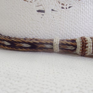 Puede incluir: Primer plano de un sombrero de vaquero blanco con una banda trenzada decorativa en colores marr&oacute;n, beige y crema. Una borla a juego cuelga de un lado. El sombrero tiene una superficie texturizada y un dise&ntilde;o occidental cl&aacute;sico.