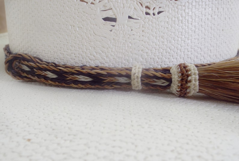 Puede incluir: Primer plano de un sombrero de vaquero blanco con una banda trenzada decorativa en colores marr&oacute;n, beige y crema. Una borla a juego cuelga de un lado. El sombrero tiene una superficie texturizada y un dise&ntilde;o occidental cl&aacute;sico.