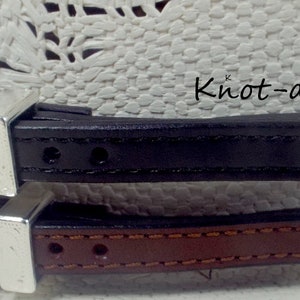 May include: Two leather hat bands with silver buckles. The top band is black and the bottom band is brown. The bands are stitched with a decorative pattern. Knot-a-Tail.com is printed on the bands.