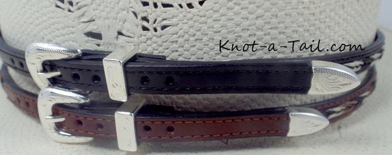 May include: Two leather hat bands with silver buckles. The top band is black and the bottom band is brown. The bands are stitched with a decorative pattern. Knot-a-Tail.com is printed on the bands.