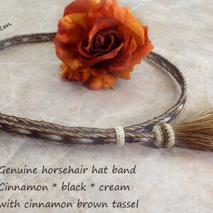 Puede incluir: Banda para sombrero de crin de caballo genuina en colores canela, negro y crema, con una borla marr&oacute;n canela. La banda est&aacute; trenzada y tiene un nudo decorativo. El texto "Knot-a-tail.cm" es visible.