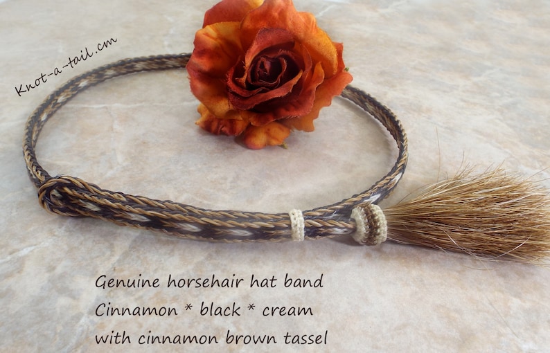 Puede incluir: Banda para sombrero de crin de caballo genuina en colores canela, negro y crema, con una borla marr&oacute;n canela. La banda est&aacute; trenzada y tiene un nudo decorativo. El texto "Knot-a-tail.cm" es visible.
