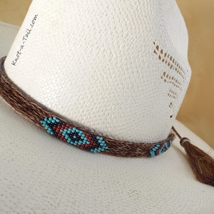 Może przedstawiać: Biały kowbojski kapelusz z brązową skórzaną opaską ozdobioną turkusowymi i brązowymi koralikami. Opaska ma mały brązowy frędzlem.