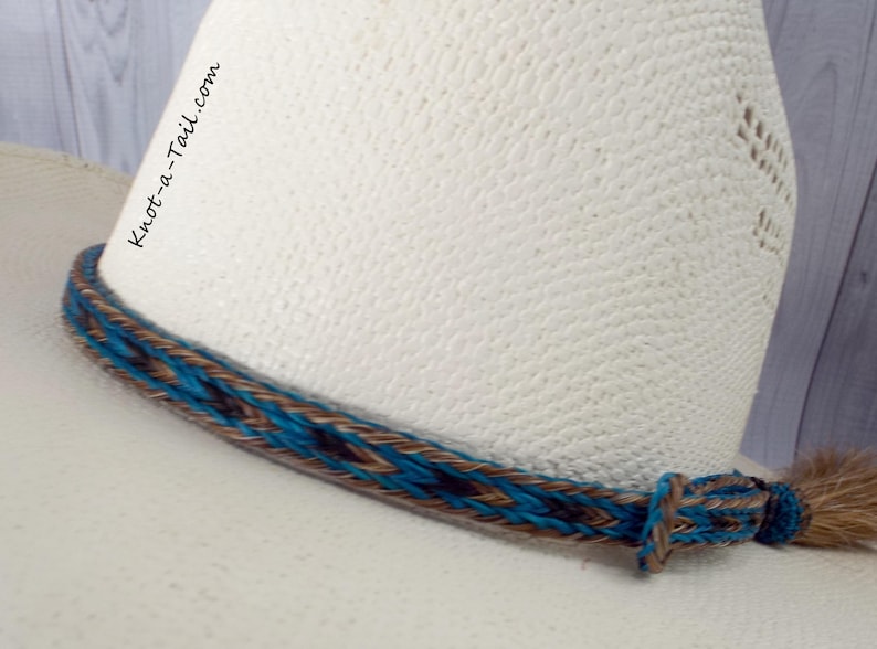 May include: A turquoise, brown, and black braided leather hat band with a long tail. The hat band is on a white cowboy hat.